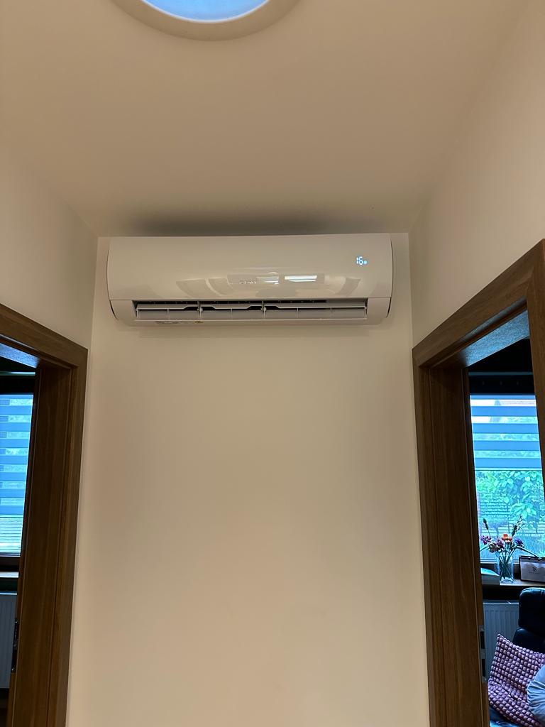 Biała jednostka klimatyzacji typu split zamontowana na ścianie w korytarzu, widoczny wyświetlacz temperatury, dwoje drzwi po bokach kadru, widok z dołu.