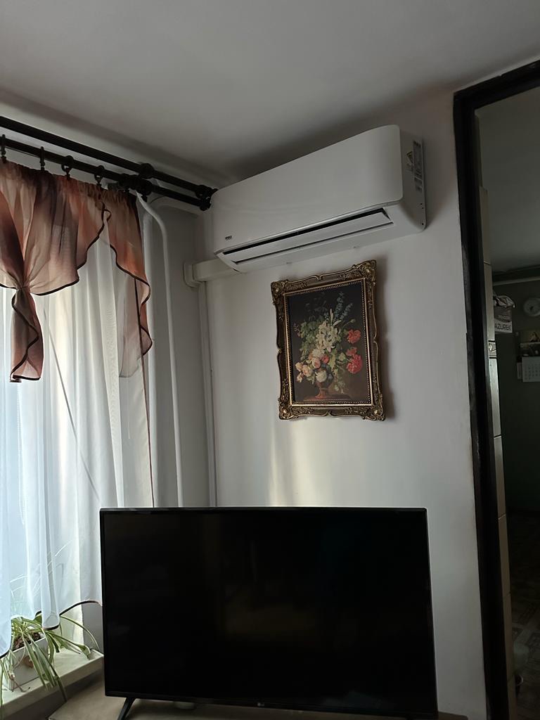 Biały klimatyzator ścienny w salonie nad czarnym telewizorem, obok okna z zasłonami i obrazem w złotej ramie.