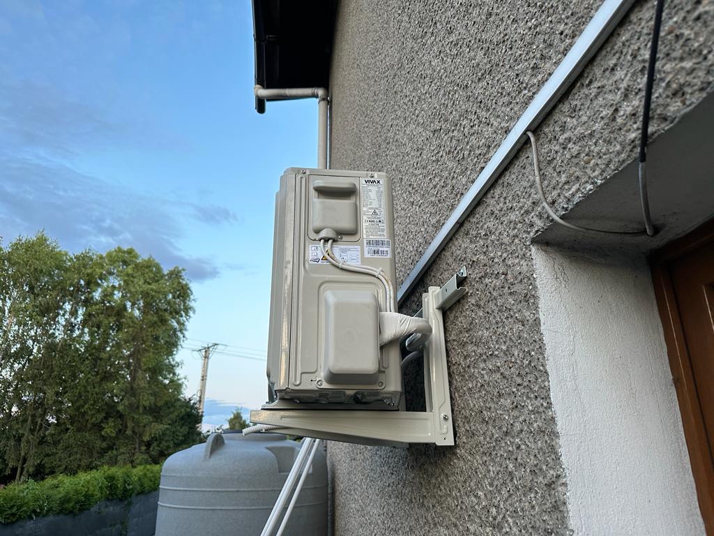Zewnętrzna jednostka klimatyzacji marki Vivax zamontowana na szarej, chropowatej elewacji domu, z widocznymi przewodami i wspornikami montażowymi, na tle zieleni i błękitnego nieba.