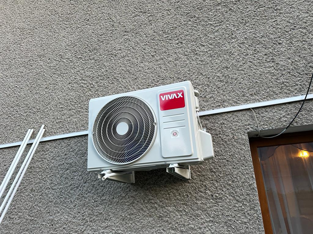 Zewnętrzna jednostka klimatyzacji VIVAX zamontowana na szarej, chropowatej ścianie budynku. Widoczne białe rury instalacyjne i fragment okna z brązową ramą po prawej stronie.