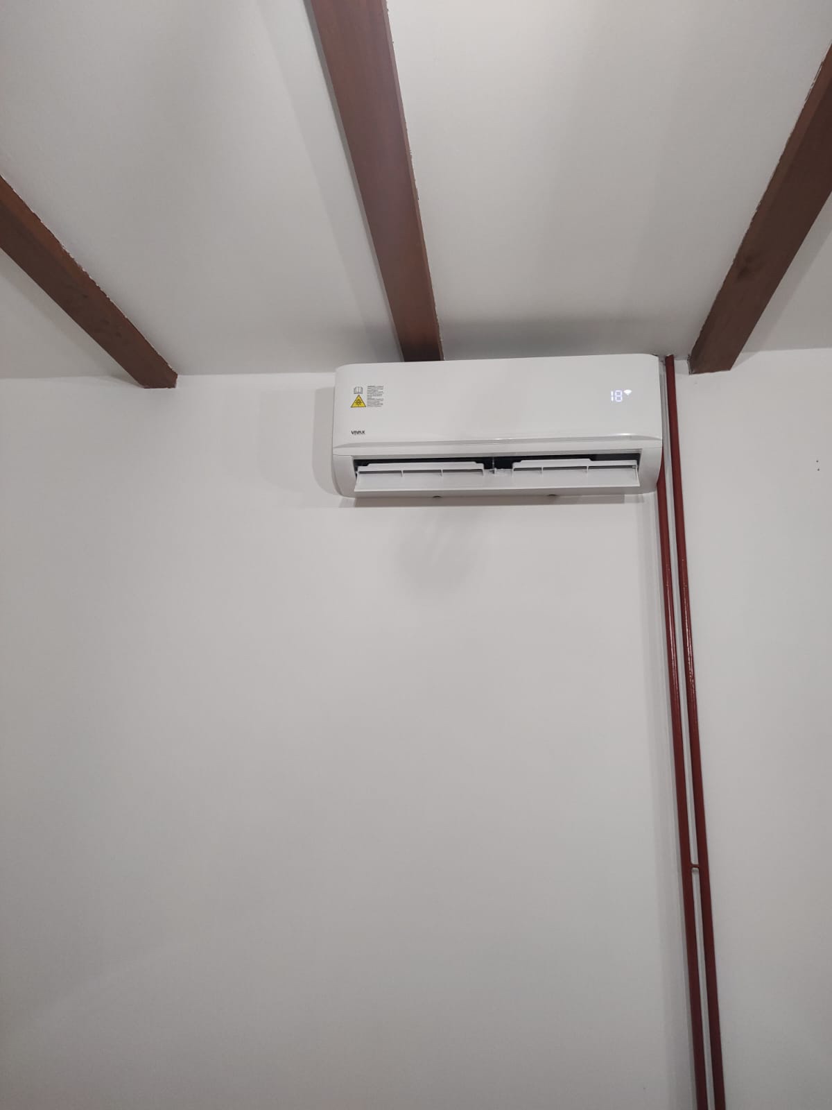 Biały klimatyzator marki Vivax zamontowany na białej ścianie w pomieszczeniu z drewnianymi belkami stropowymi i pionową instalacją rurową w kolorze czerwonym.