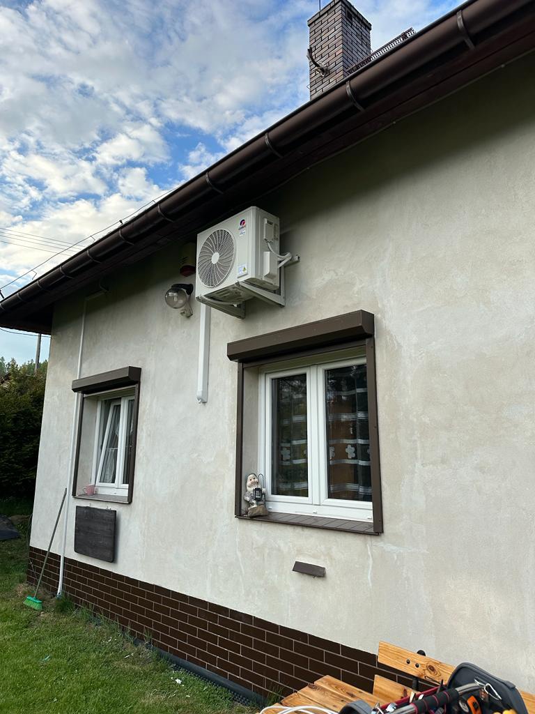 Zewnętrzna jednostka klimatyzacji zamontowana na elewacji domu z brązowym dachem i oknami z roletami, widok z poziomu trawnika.