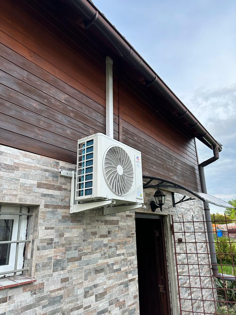 Biała jednostka zewnętrzna klimatyzatora zamontowana na elewacji domu z brązowymi drewnianymi panelami i kamienną okładziną, widoczny przewód odprowadzający skropliny.