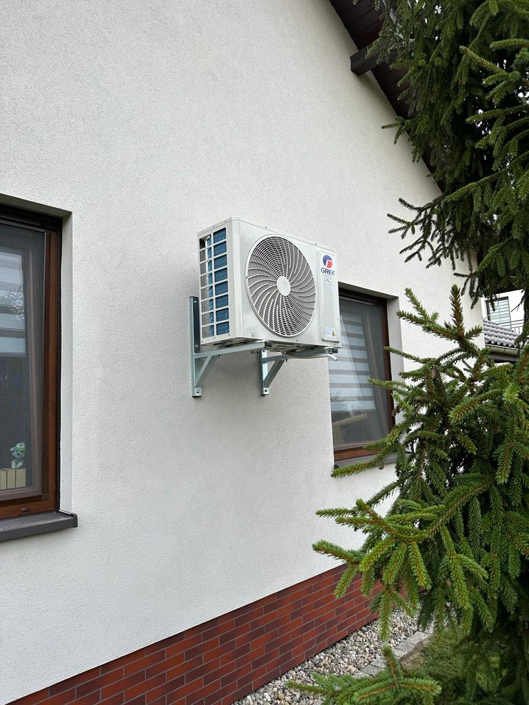 Zewnętrzna jednostka klimatyzacji marki Gree zamontowana na białej elewacji domu z ceglaną podmurówką, widoczna z boku, z fragmentem okna i iglaka w kadrze.