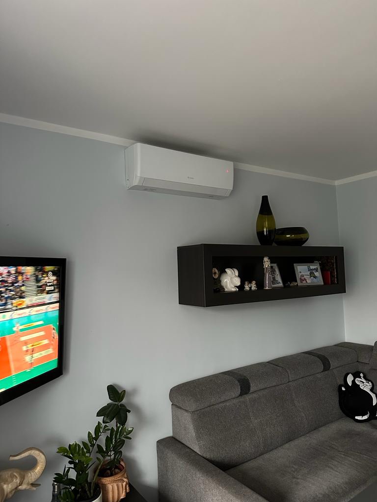 Biała jednostka klimatyzacyjna zamontowana na ścianie w salonie, widoczny telewizor z włączonym meczem siatkówki oraz szara sofa.