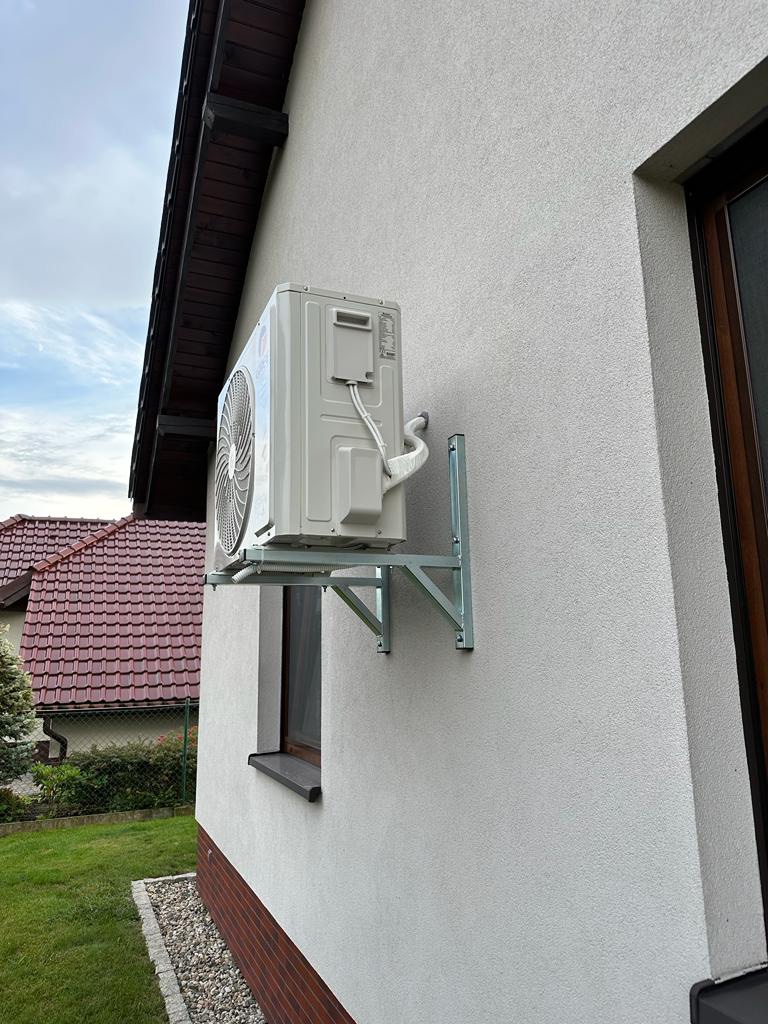 Zewnętrzna jednostka klimatyzacji zamontowana na ścianie domu, widok z dołu, z widocznymi elementami montażowymi i fragmentem elewacji oraz dachu sąsiedniego budynku.