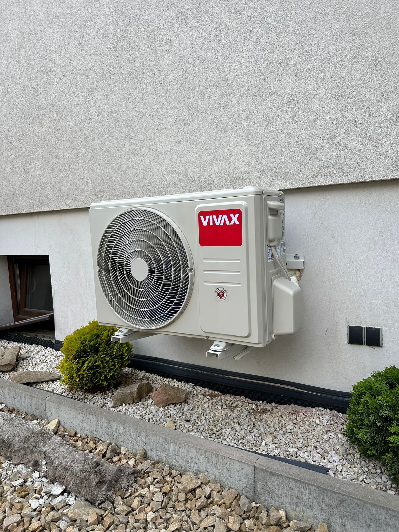 Zewnętrzna jednostka klimatyzacji VIVAX zamontowana na elewacji domu, widok z poziomu ogrodu z kamienną rabatą i krzewami.