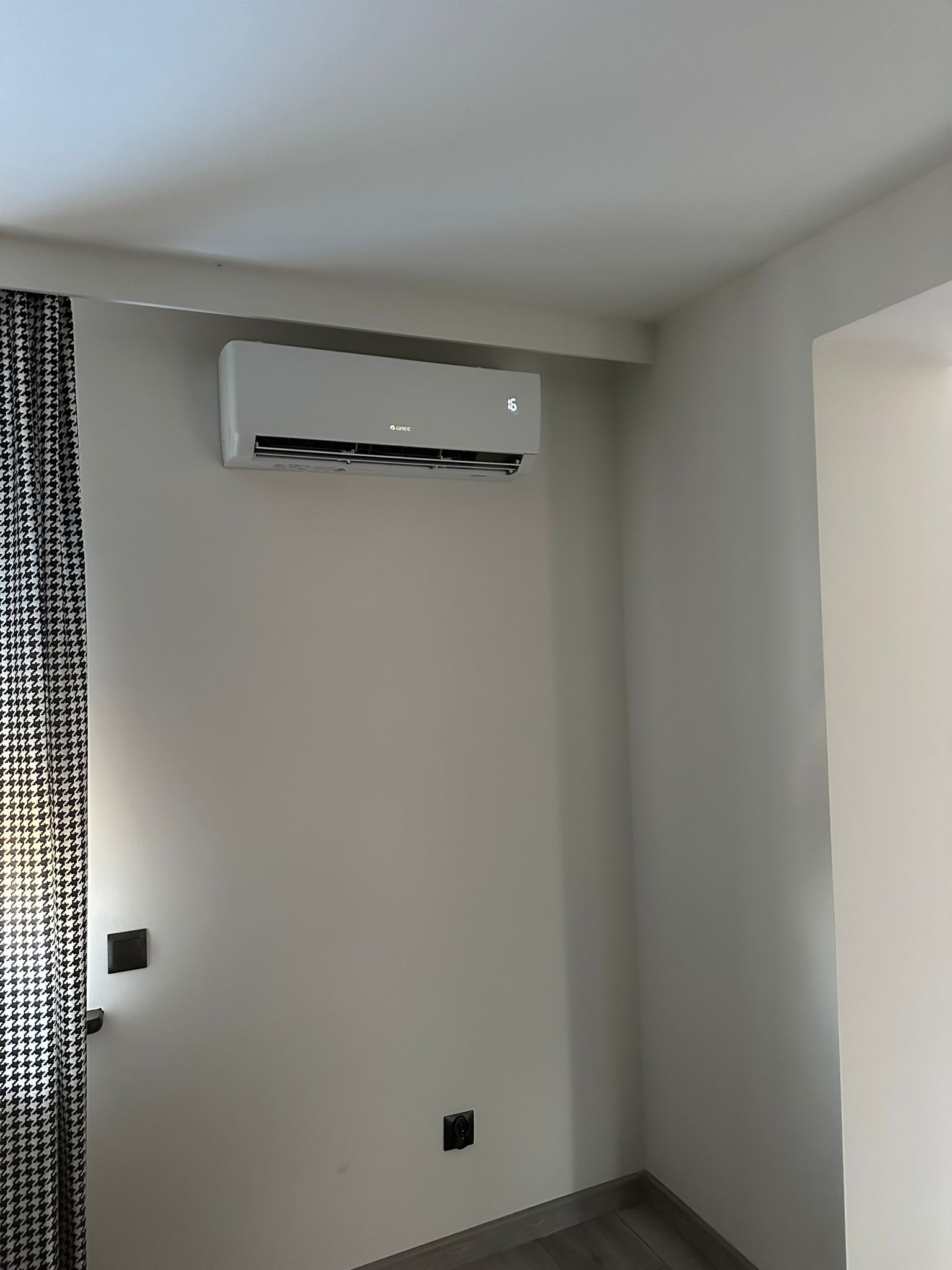 Biały klimatyzator marki Gree zamontowany na ścianie w pokoju z zasłoną w czarno-białą pepitkę i ciemnymi gniazdkami.