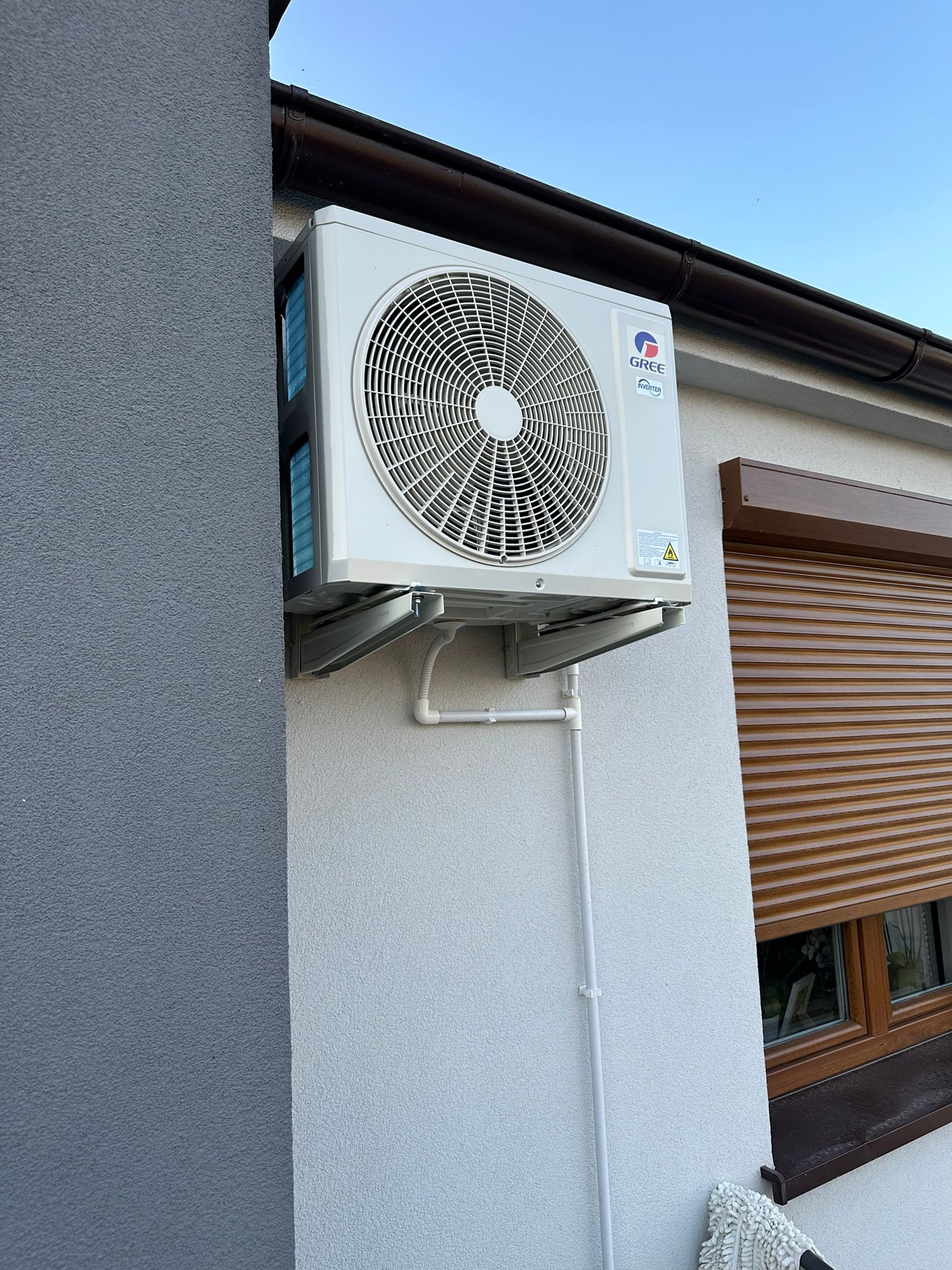 Zewnętrzna jednostka klimatyzacji marki Gree zamontowana na elewacji budynku, widoczne rury odprowadzające skropliny, z prawej strony okno z brązowymi roletami zewnętrznymi.