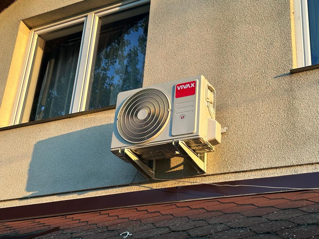 Zewnętrzna jednostka klimatyzacji VIVAX zamontowana na elewacji budynku, widoczna nad brązowym dachem, oświetlona ciepłym światłem zachodzącego słońca.