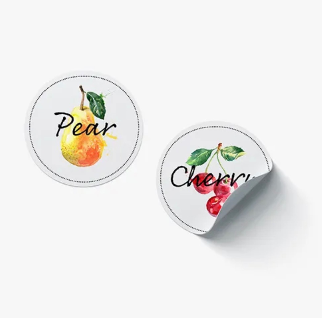 Dwa okrągłe, białe arkusze etykiet z grafikami akwarelowymi: gruszka z napisem 'Pear' i wiśnie z napisem 'Cherry', jeden arkusz częściowo odklejony, rzucający cień.