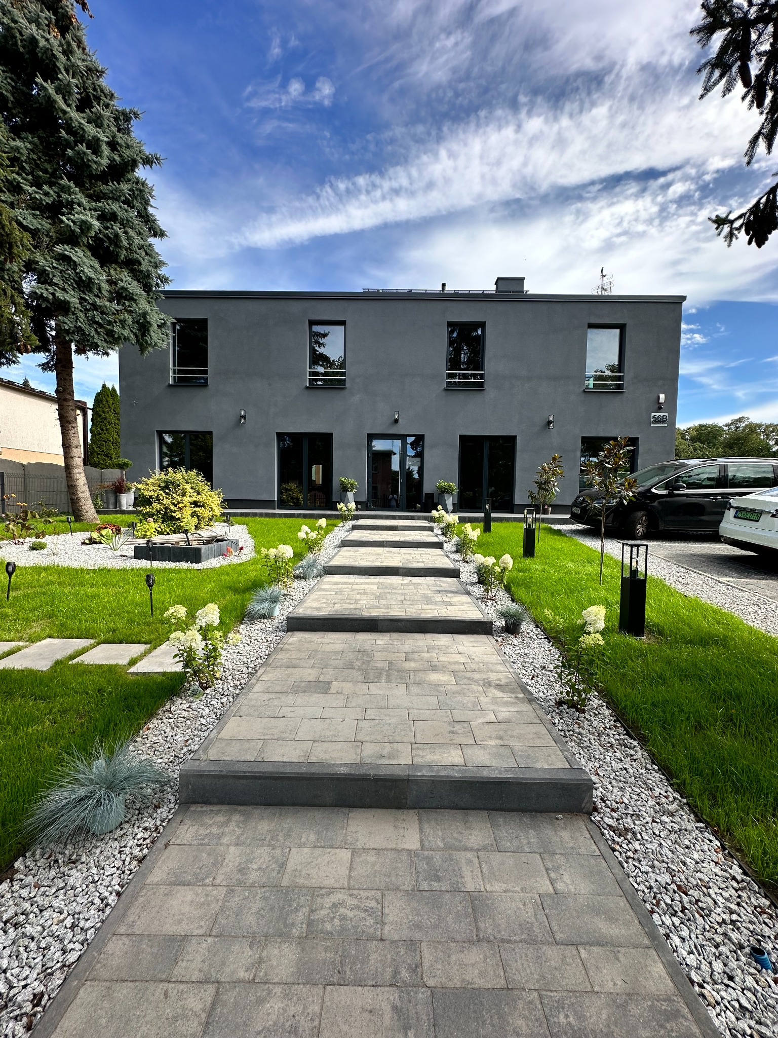 Nowoczesny dom jednorodzinny z szarą elewacją, brukowaną ścieżką prowadzącą do wejścia, zadbanym trawnikiem, rabatami z białymi kwiatami i ozdobnymi kamieniami, widok z perspektywy ścieżki.