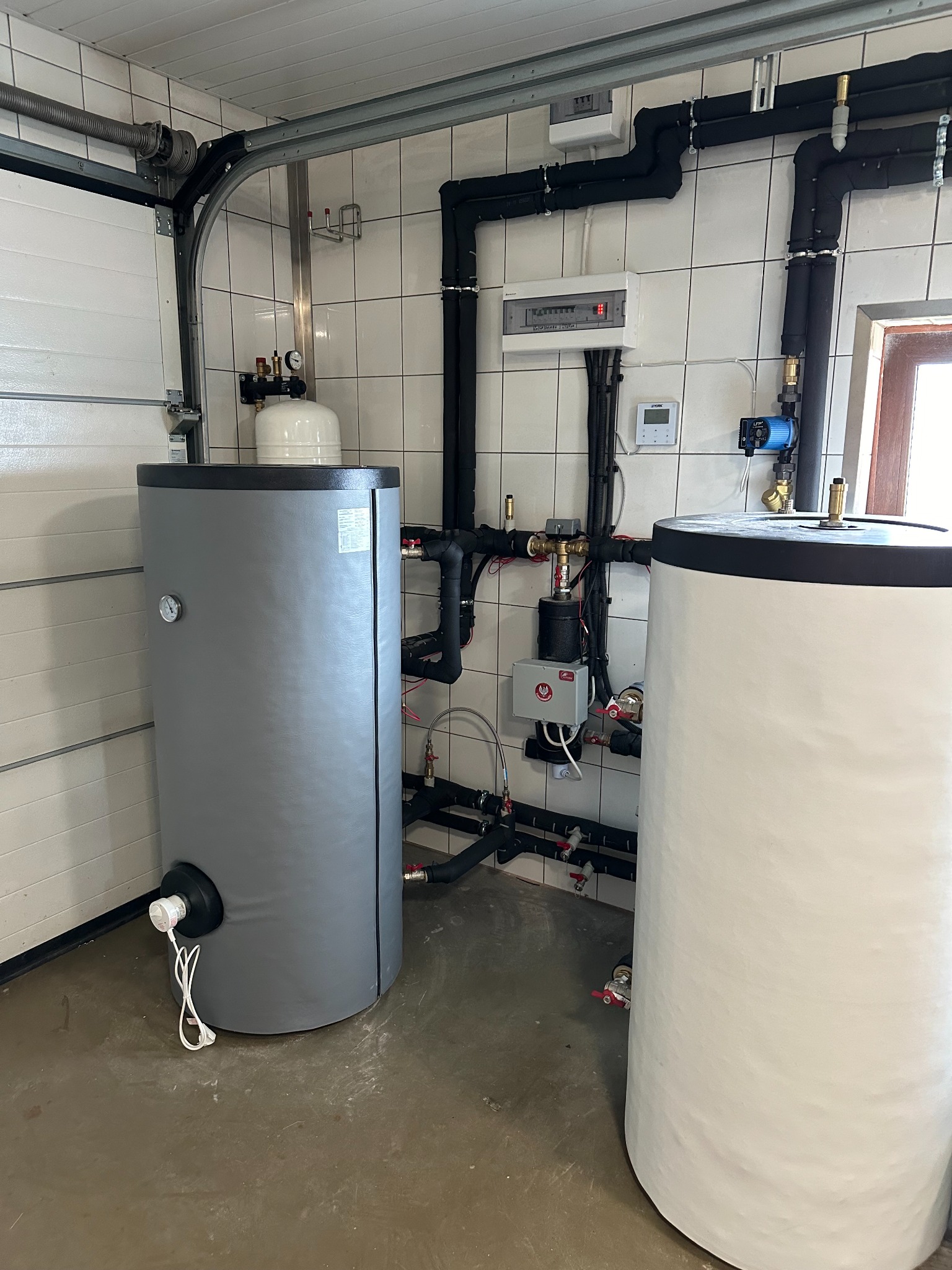 Dwa zbiorniki akumulacyjne ciepłej wody w kotłowni w Szczecinie, z widocznymi rurami, zaworami i elementami sterującymi na ścianie z białych płytek.