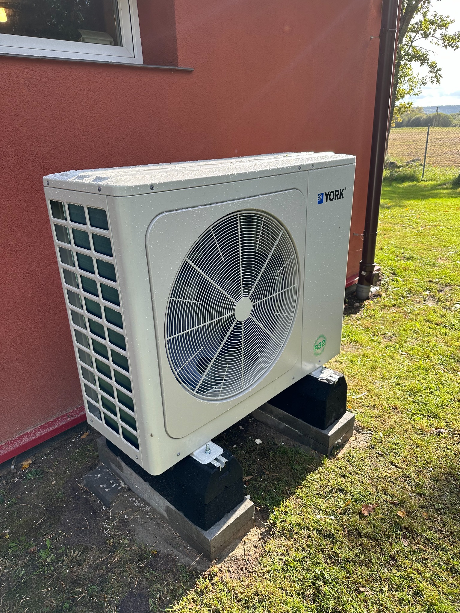 Biała jednostka zewnętrzna klimatyzacji YORK, z kroplami wody na obudowie, zamontowana na betonowych podstawach przy ścianie budynku o czerwonej elewacji, widok na zielony trawnik i pole w tle.