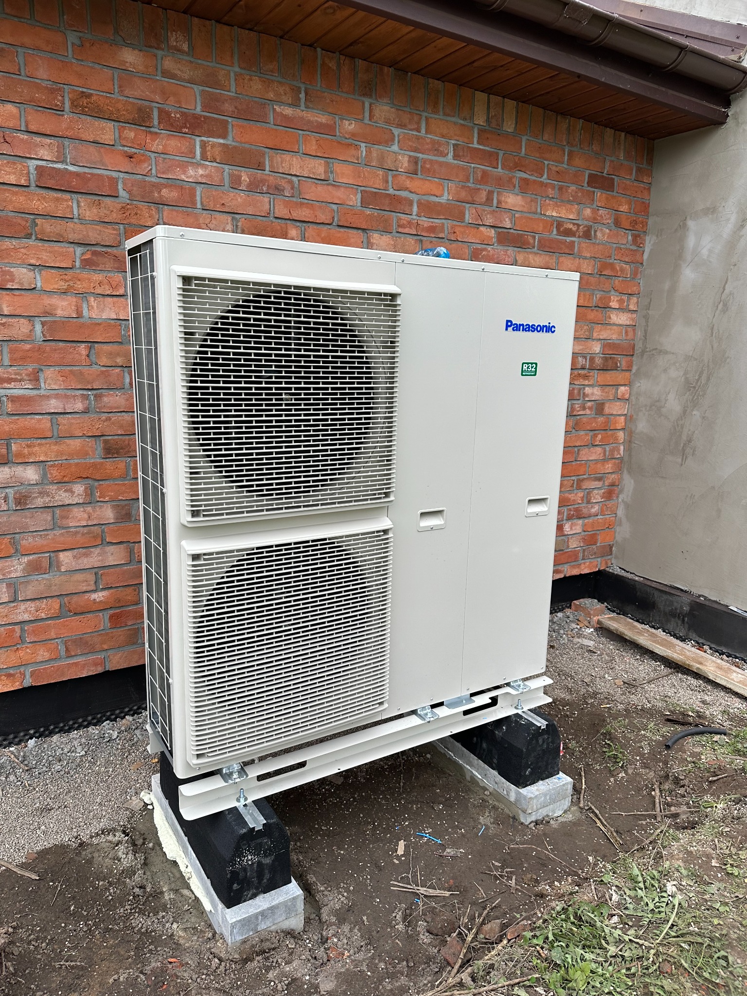 Zewnętrzna jednostka pompy ciepła Panasonic zamontowana na betonowych podstawach z izolacją, na tle ceglanej ściany budynku.