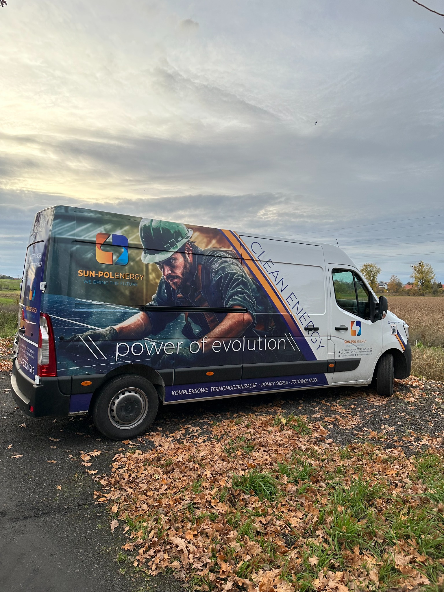 Biały samochód dostawczy z grafiką firmy zajmującej się odnawialnymi źródłami energii, zaparkowany na poboczu drogi pokrytej liśćmi. Grafika na samochodzie przedstawia instalatora w kasku...