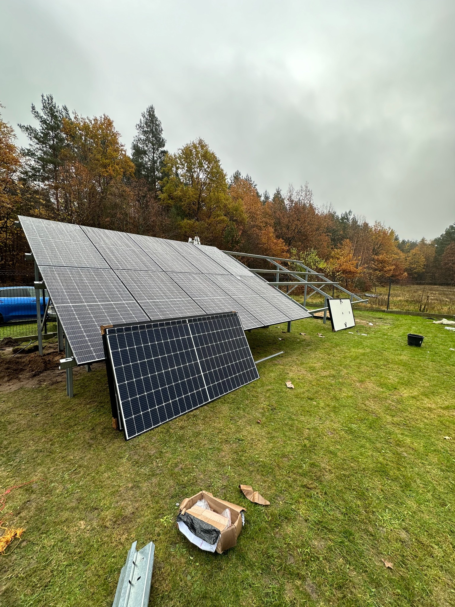 Panele słoneczne w trakcie montażu na zielonej trawie, widoczna konstrukcja wsporcza i kartonowe pudło po elementach, jesienne drzewa w tle, pochmurne niebo.