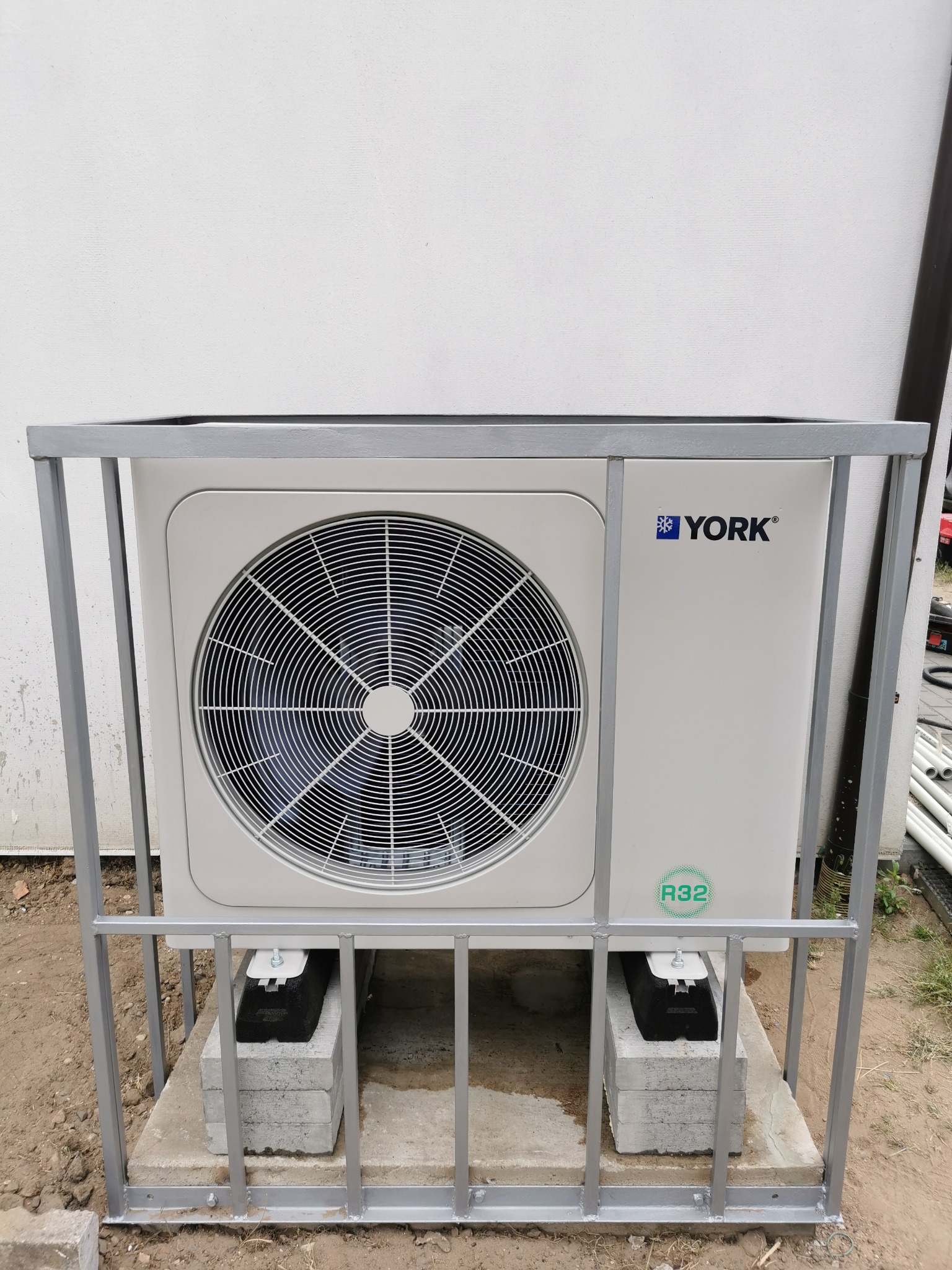 Zewnętrzna jednostka pompy ciepła marki York, umieszczona na betonowych bloczkach i zabezpieczona metalową klatką, widoczny wentylator i oznaczenie R32.