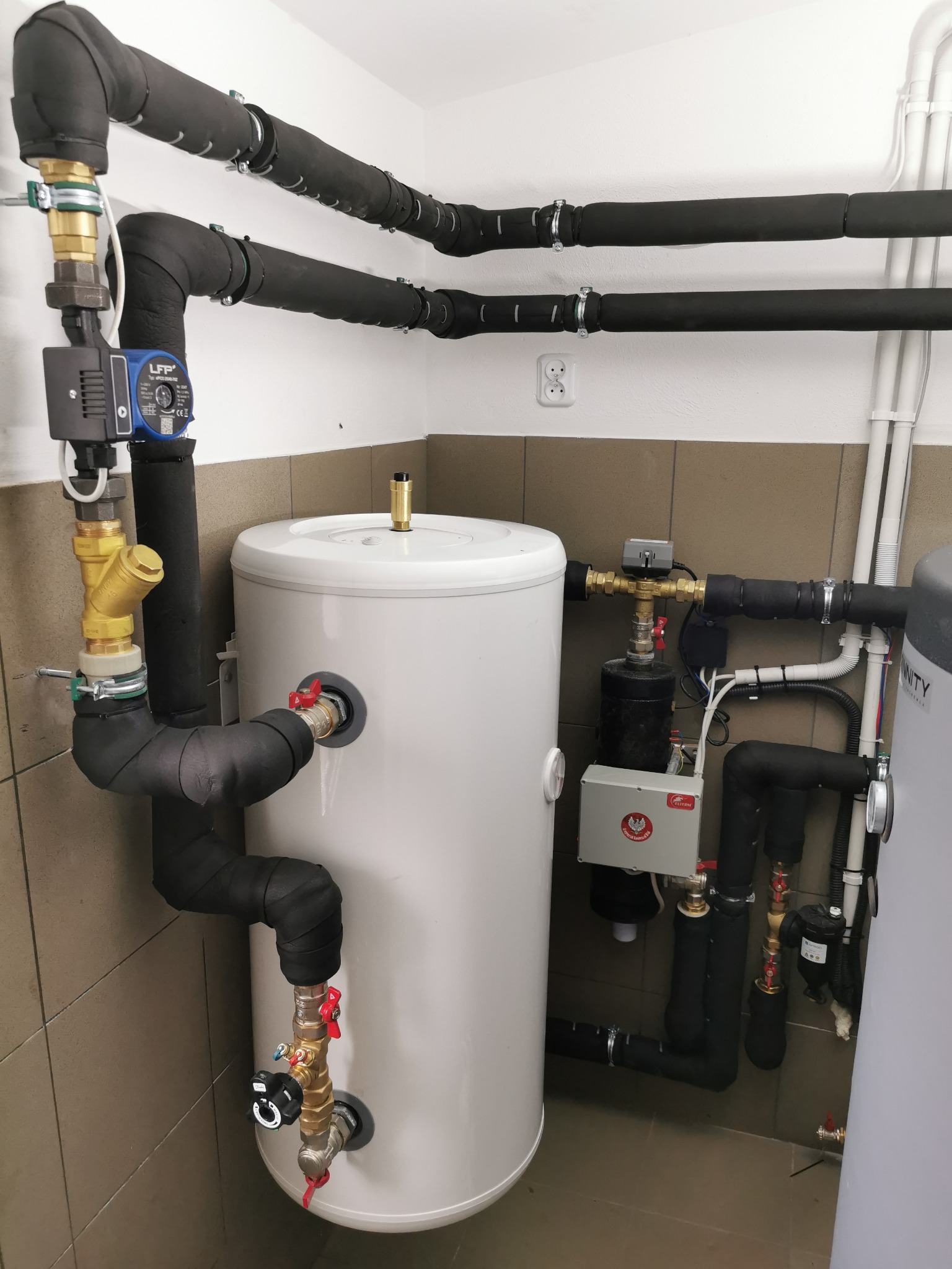 Biały zasobnik ciepłej wody użytkowej z czarnymi, zaizolowanymi rurami, zaworami i pompą obiegową w pomieszczeniu z kafelkami na ścianach.