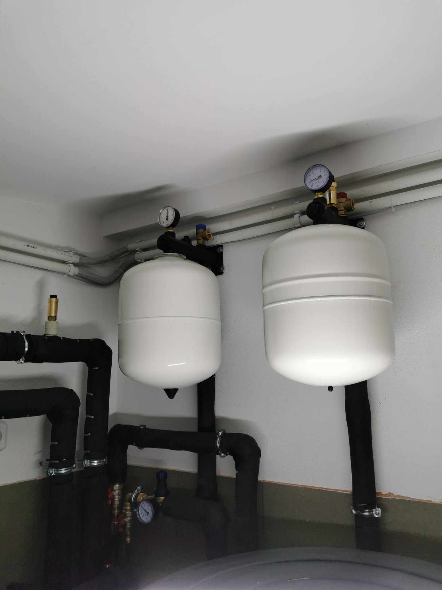 Dwa białe zbiorniki wyrównawcze ciśnienia z manometrami, podłączone czarnymi, izolowanymi rurami, zamocowane na ścianie w kotłowni.