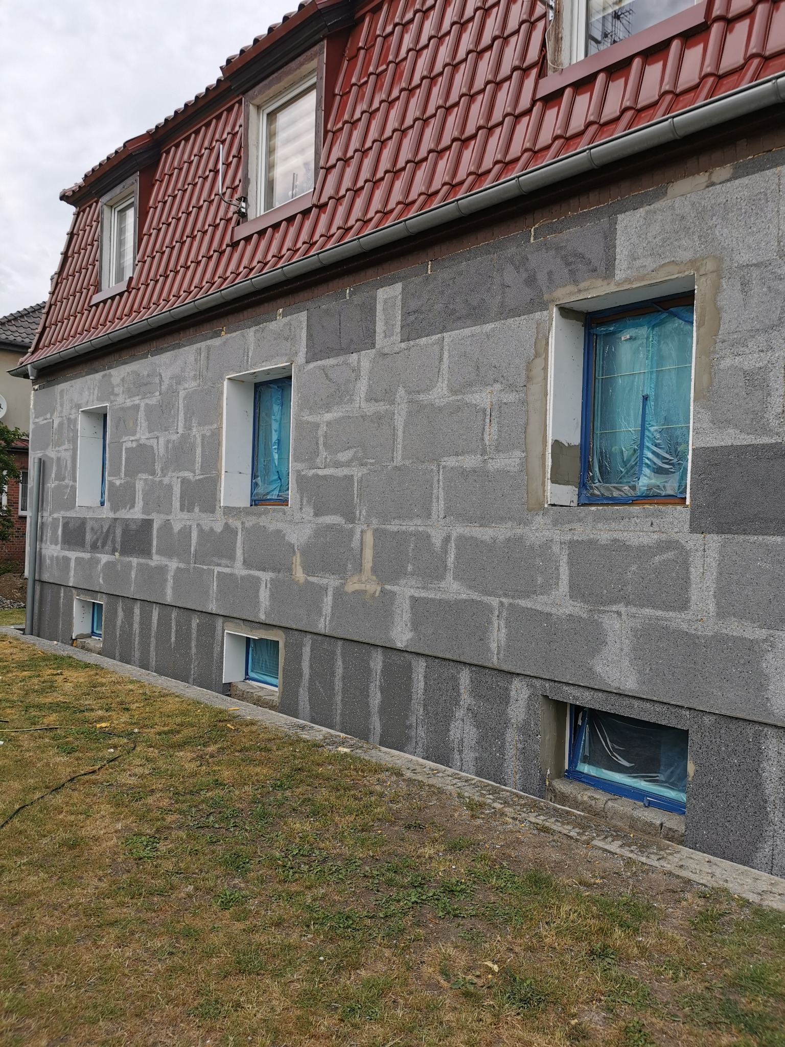Dom w trakcie ocieplania: szare bloczki na ścianie, okna zabezpieczone folią, widoczny fragment dachu z czerwoną dachówką.