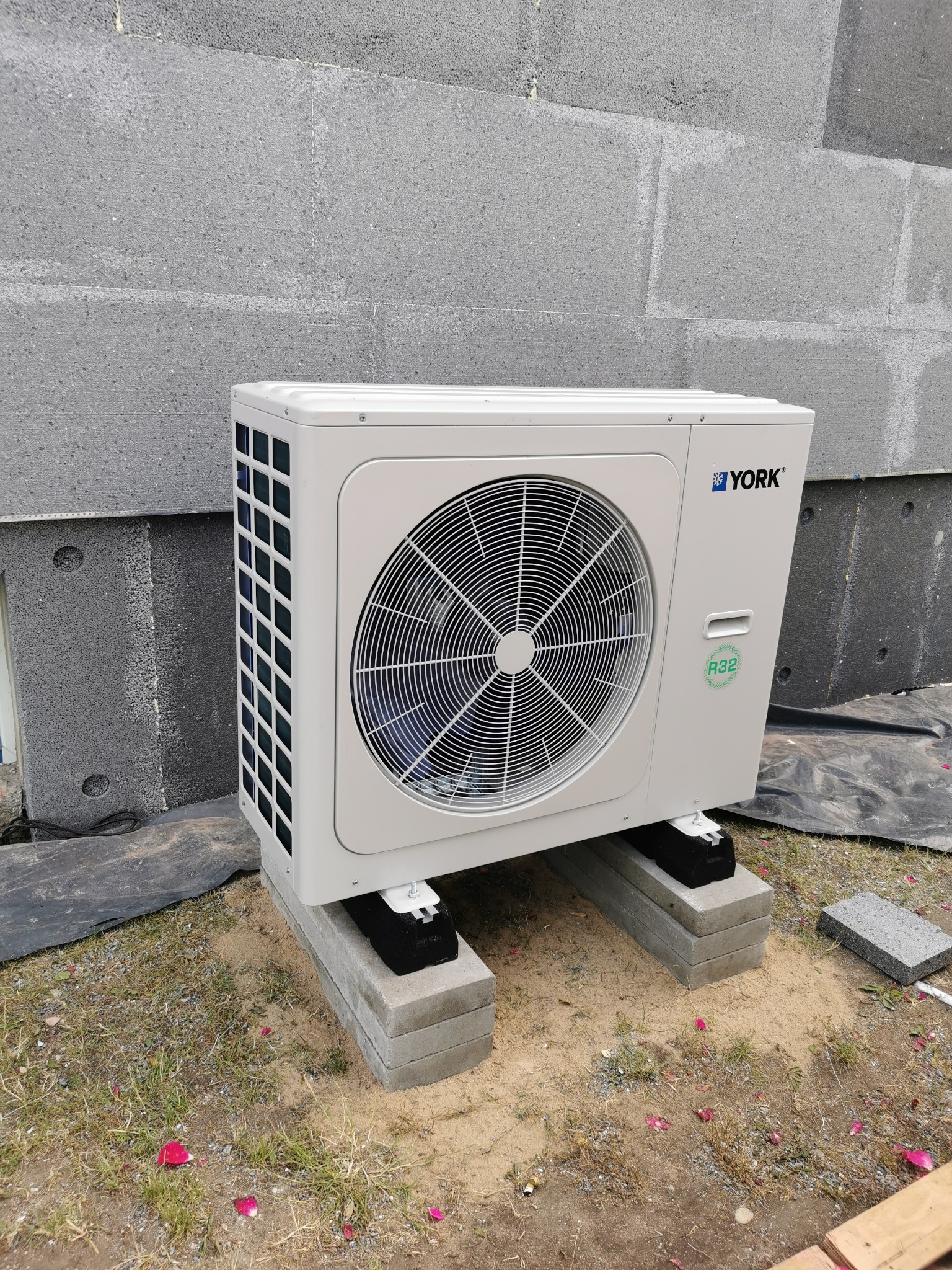 Biała jednostka zewnętrzna pompy ciepła marki York, umieszczona na betonowych bloczkach, z widocznym logo i oznaczeniem czynnika chłodniczego R32, na tle szarej ściany budynku z betonu komórkowego.