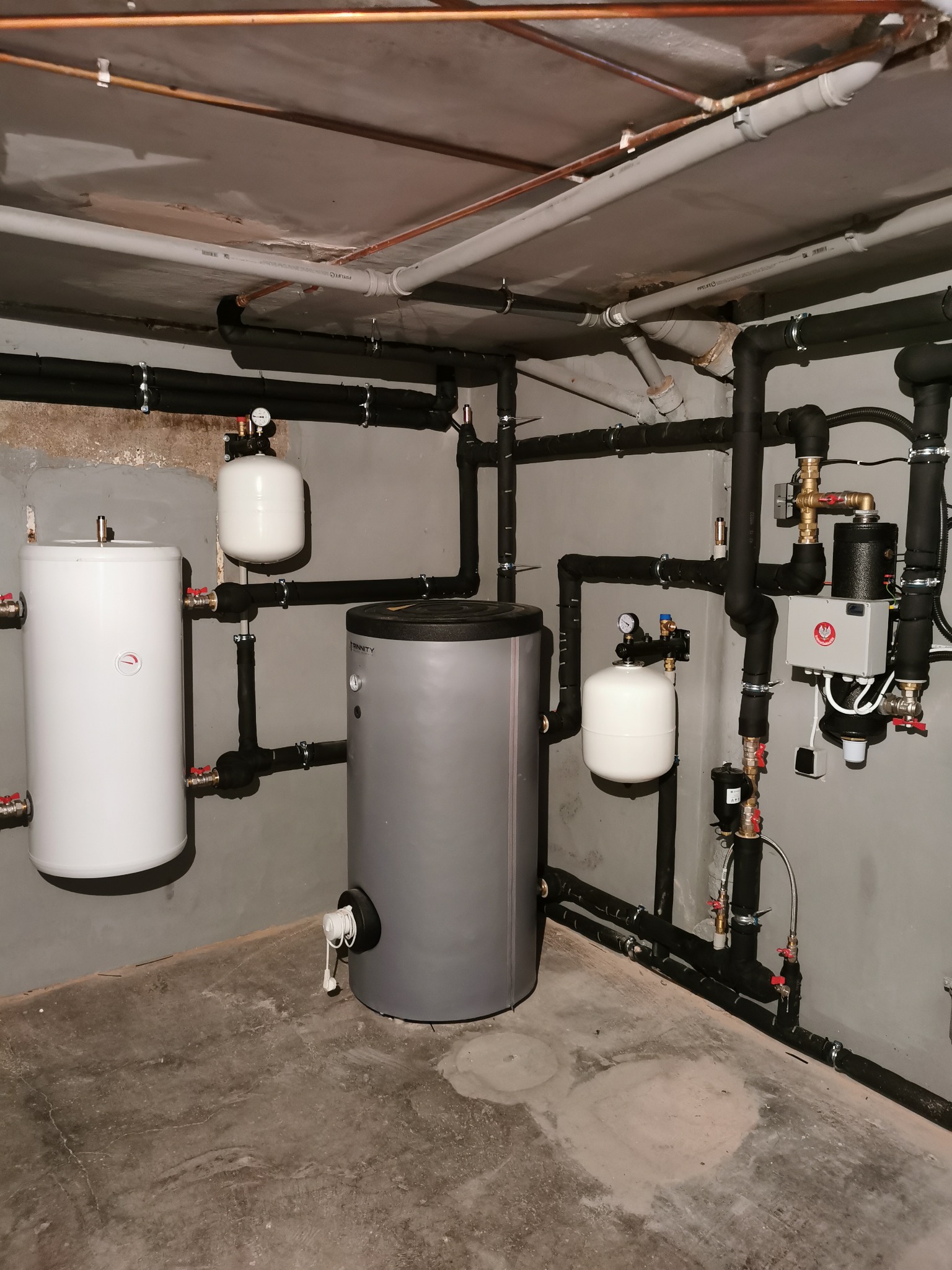 Instalacja w kotłowni z dwoma zbiornikami akumulacyjnymi, białym i szarym, połączonymi systemem izolowanych rur i zaworów, z widocznymi manometrami i elementami sterującymi. Rury prowadzone...