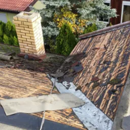 Uszkodzony dach z częściowo zerwanym pokryciem, widoczne fragmenty papy i blachy, ceglany komin w tle, uszkodzenia spowodowane warunkami atmosferycznymi.