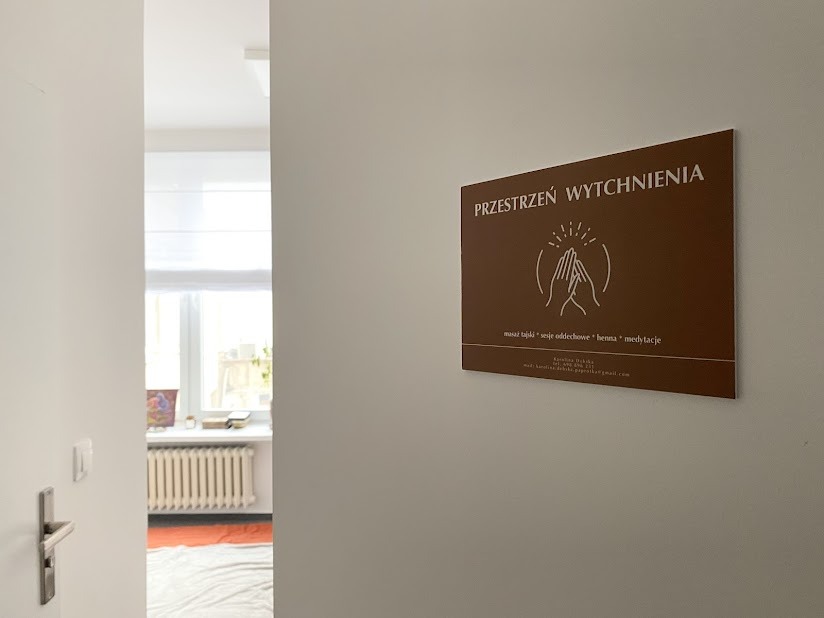 Brązowa tabliczka informacyjna z napisem 'Przestrzeń Wytchnienia' i grafiką dłoni, umieszczona na białej ścianie obok otwartych drzwi do jasnego pokoju z grzejnikiem i oknem.