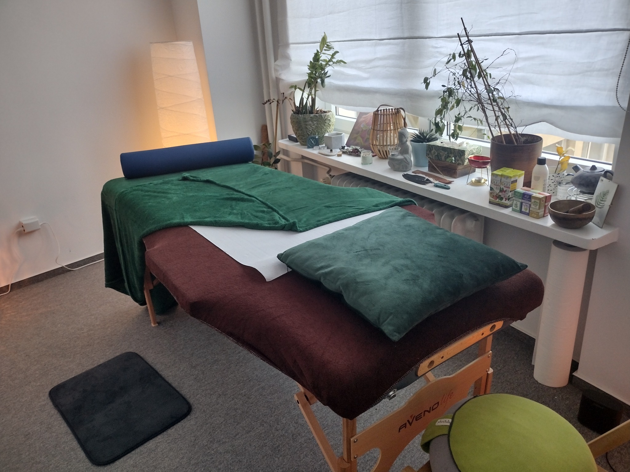 Przygotowany stół do zabiegów z zielonym pledem i poduszką, wałek pod głowę. Na parapecie okiennym rośliny doniczkowe i dekoracje. W tle lampa podłogowa.