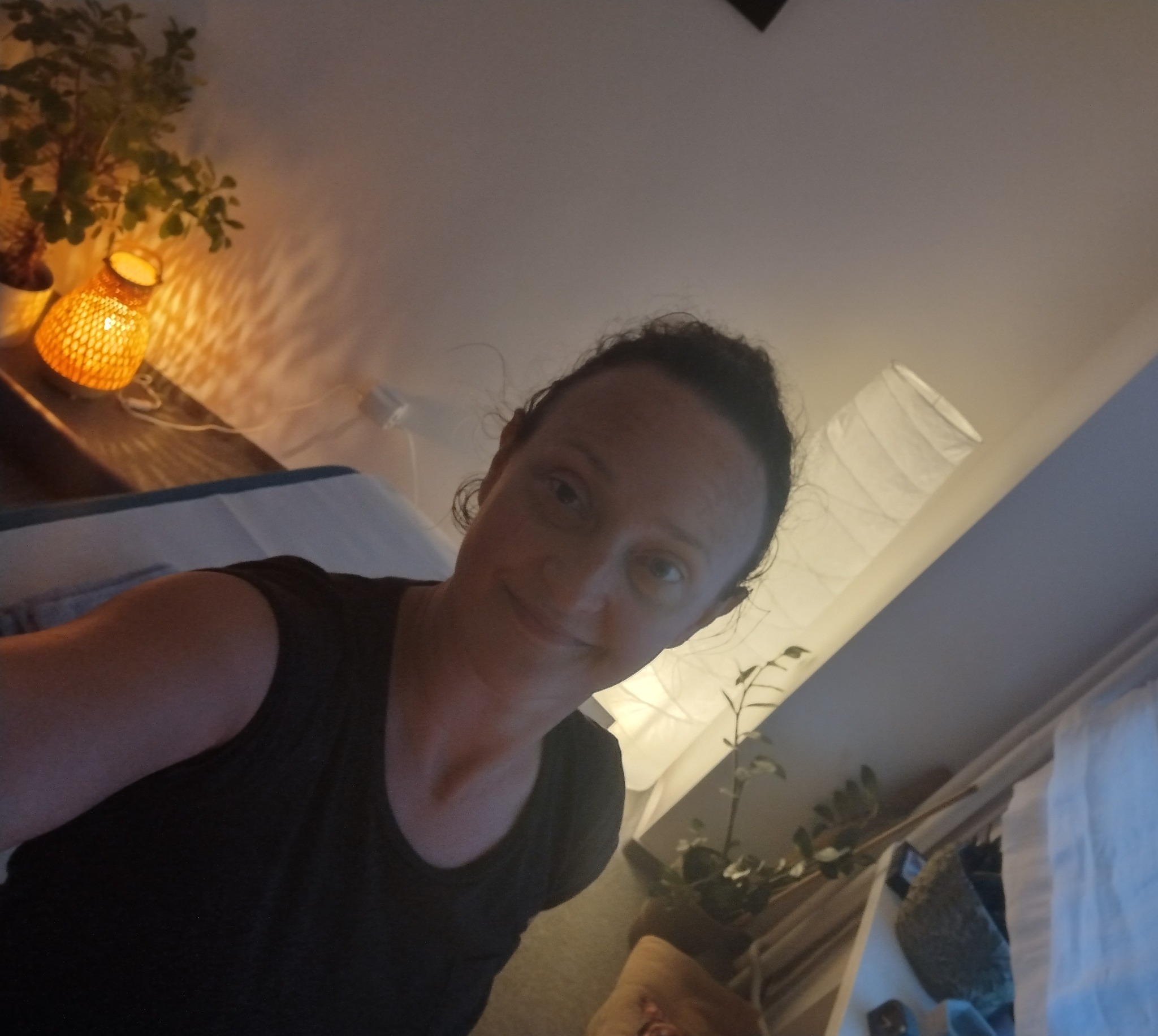 Kobieta robiąca sobie selfie w pokoju z łóżkiem do masażu, obok lampka z wikliny i roślina w doniczce.