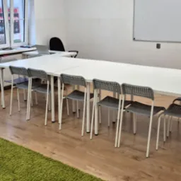 Sala lekcyjna z długim białym stołem, szarymi krzesłami i zielonym dywanem z logo szkoły językowej w rogu. Na ścianie tablica suchościeralna i zegar.