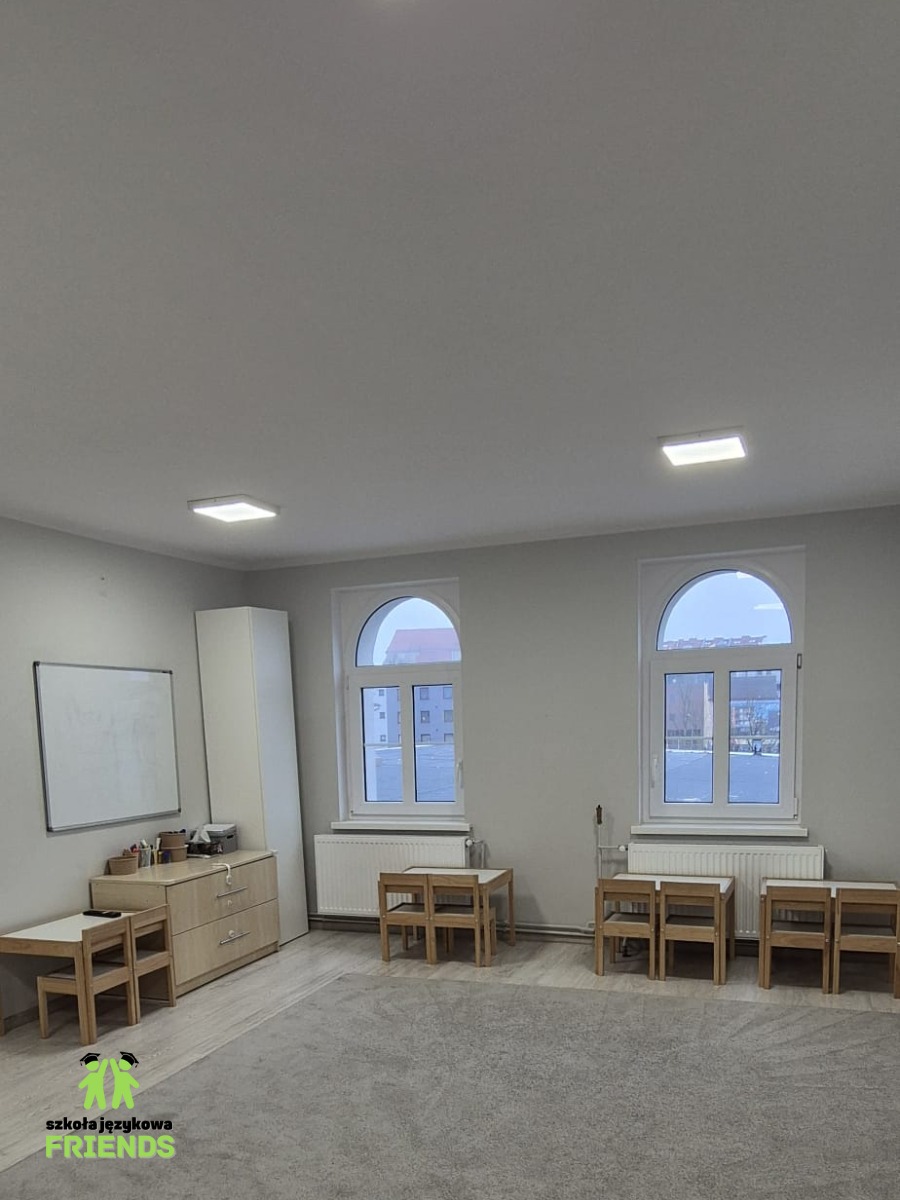 Pusta sala lekcyjna z dwoma oknami łukowymi, białymi ścianami, biurkiem, szafką, tablicą i zestawami małych drewnianych stolików z krzesłami, przygotowana do zajęć.
