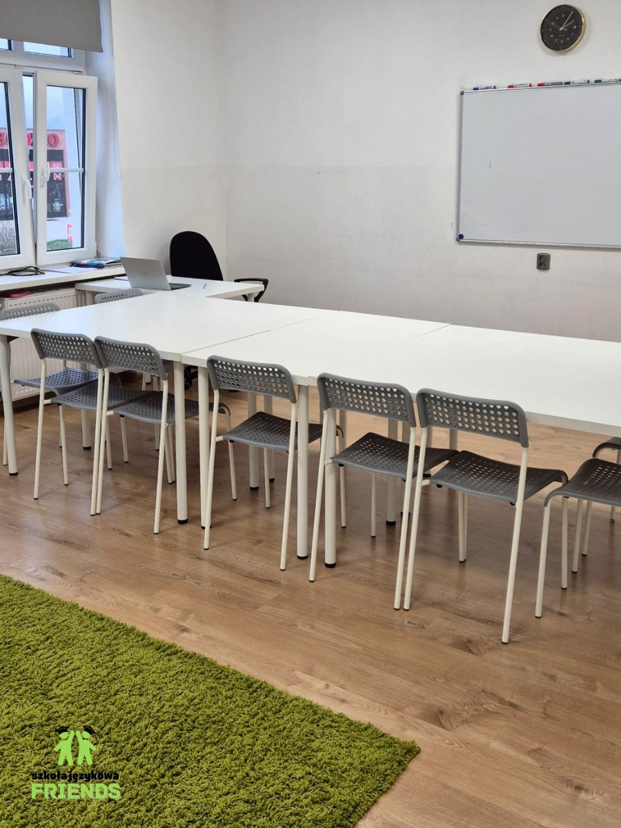 Sala lekcyjna z długim białym stołem, szarymi krzesłami i zielonym dywanem z logo szkoły językowej w rogu. Na ścianie tablica suchościeralna i zegar.