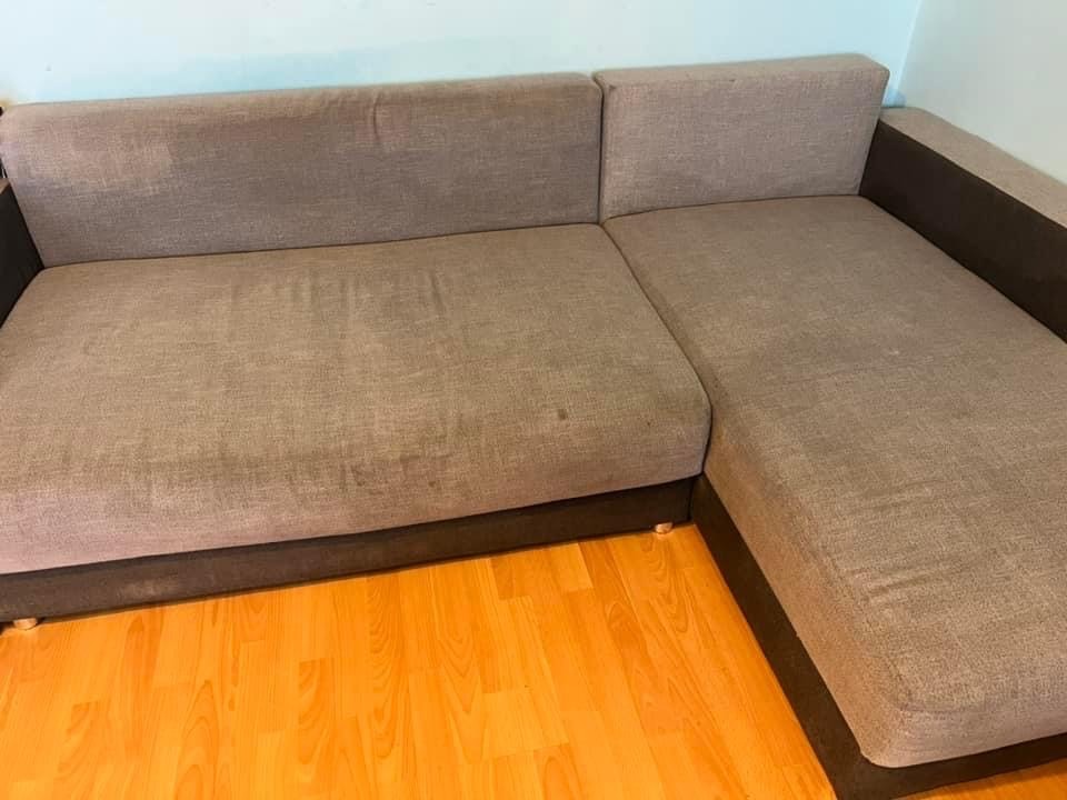 Szara narożna kanapa z widocznymi plamami na tapicerce, stojąca na parkiecie w słonecznym świetle.