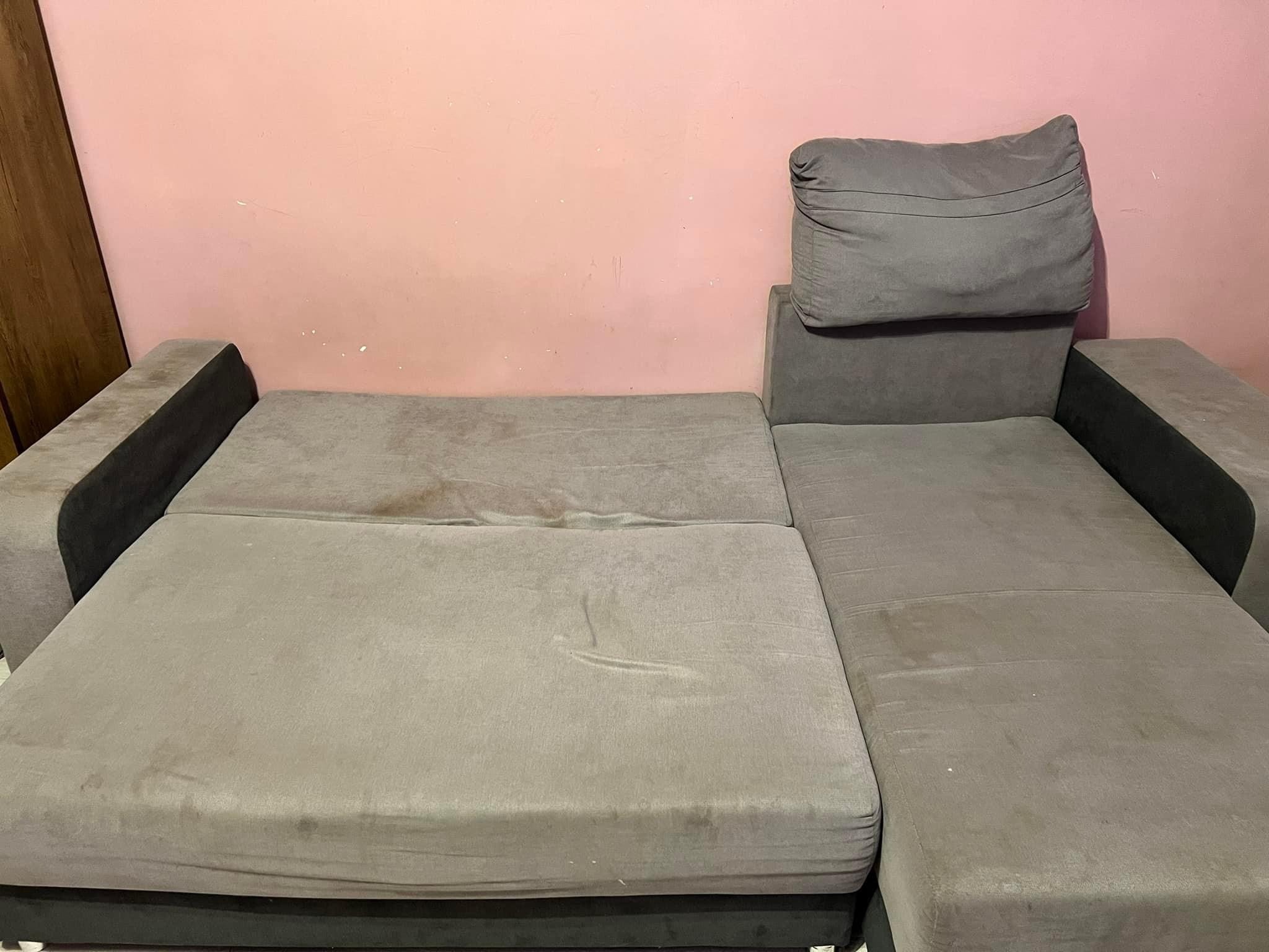 Szara, rozłożona kanapa z widocznymi plamami na obiciu i śladami użytkowania, stojąca na tle różowej ściany.