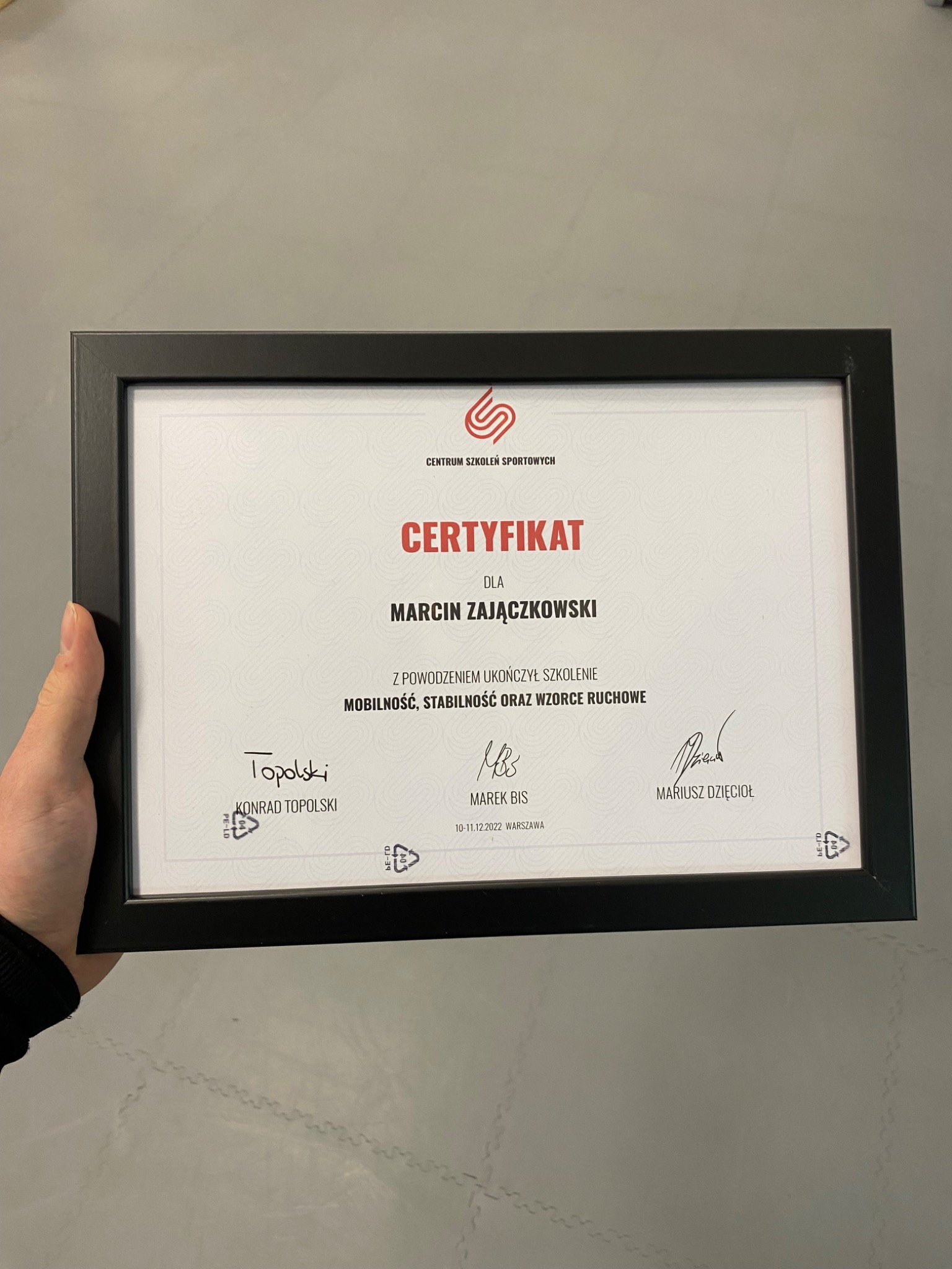 Czarno-biała ramka z certyfikatem ukończenia szkolenia z mobilności, stabilności i wzorców ruchowych dla Marcina Zajączkowskiego, trzymana w ręce na szarym tle.