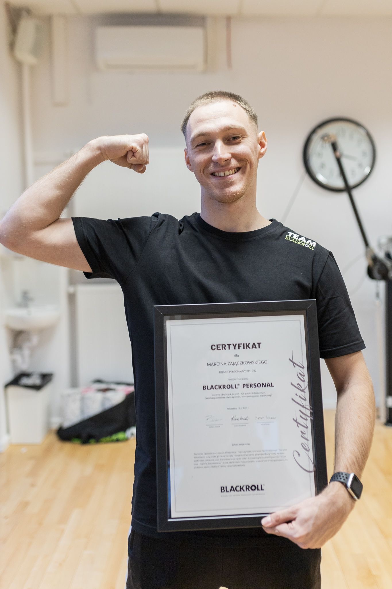 Uśmiechnięty trener personalny z Lublina prezentuje certyfikat Blackroll Personal, unosząc z dumą umięsień biceps; w tle widoczny zegar ścienny.