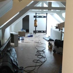 Usługi budowlano remontowe - Poddasze w trakcie prac wykończeniowych, widoczne białe belki stropowe, ściany przygotowane do malowania, sprzęt budowlany i kable na podłodze.