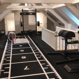 Usługi budowlano remontowe - Montaż czarnej, gumowej podłogi w domowej siłowni na poddaszu z numeracją od 1 do 9; widoczny sprzęt sportowy i białe, drewniane belki stropowe.