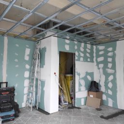 Usługi budowlano remontowe - Wnętrze w trakcie prac wykończeniowych: widoczne metalowe profile sufitowe, częściowo oszlifowane ściany z nałożoną gładzią, drabina oparta o ścianę, skrzynki narzędziowe i materiały budowlane...