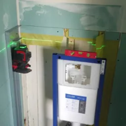 Stelaż podtynkowy Cersanit zamontowany w narożniku łazienki, widoczny laser krzyżowy, ściany częściowo obłożone płytą kartonowo-gipsową, poziomica na stelażu.