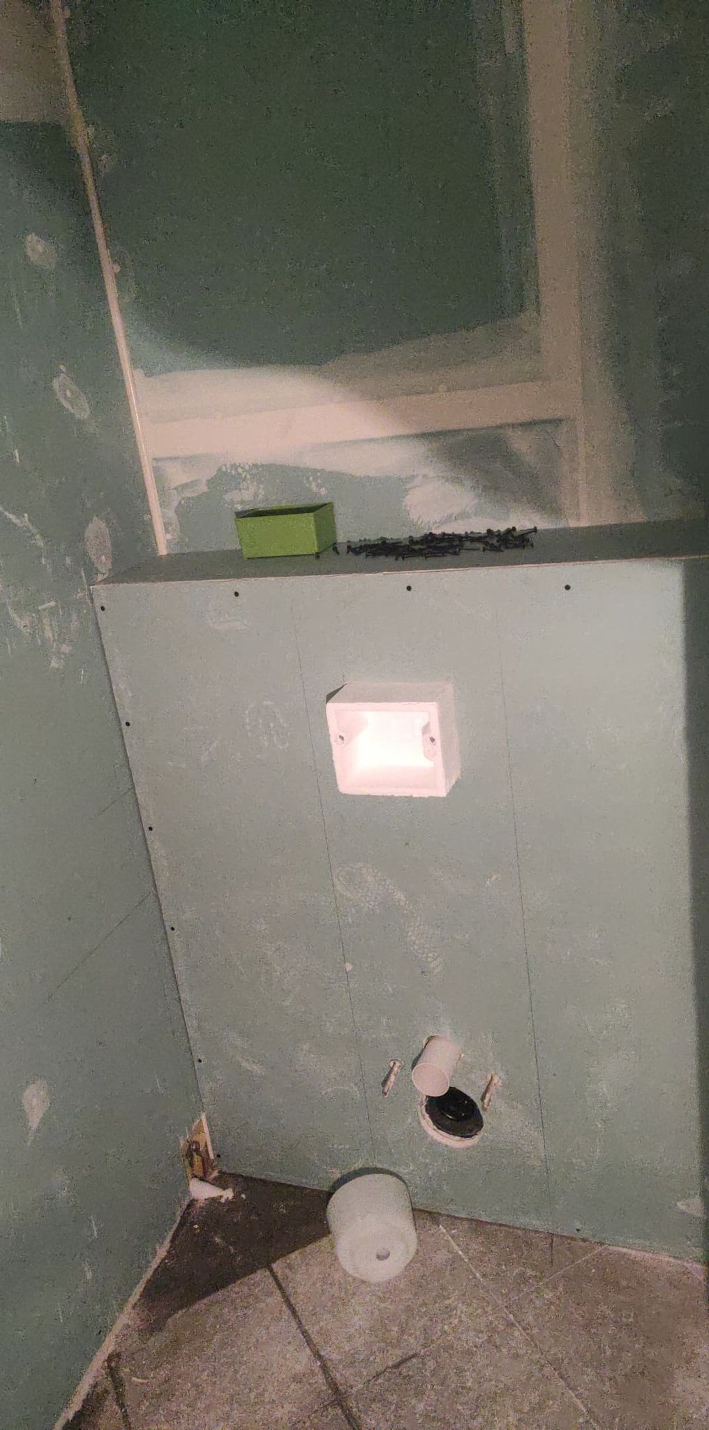 Niedokończona łazienka: ściany z płyt kartonowo-gipsowych, widoczna puszka elektryczna, otwór kanalizacyjny i rolka papieru toaletowego na brudnej podłodze z płytek.
