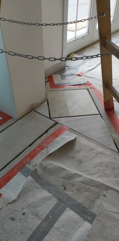 Przygotowanie podłoża pod nową podłogę: zabezpieczone folią i taśmą podłoże z widocznymi szczelinami między płytami, drabina w rogu kadru, łańcuch zabezpieczający obszar robót.