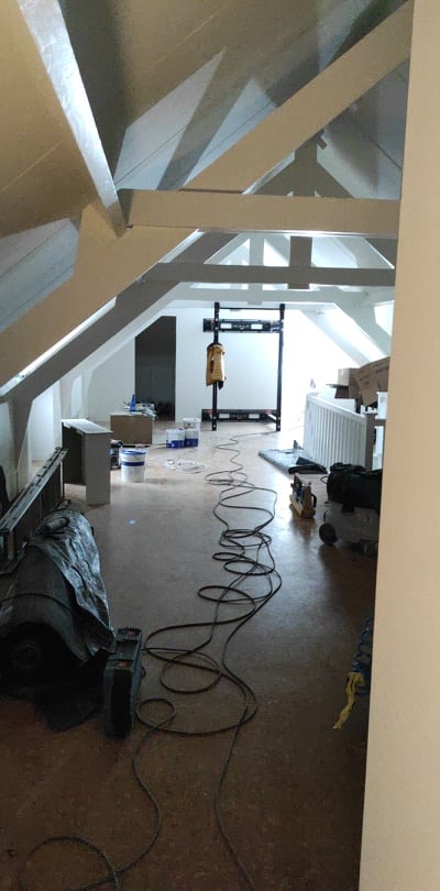 Poddasze w trakcie prac wykończeniowych, widoczne białe belki stropowe, ściany przygotowane do malowania, sprzęt budowlany i kable na podłodze.