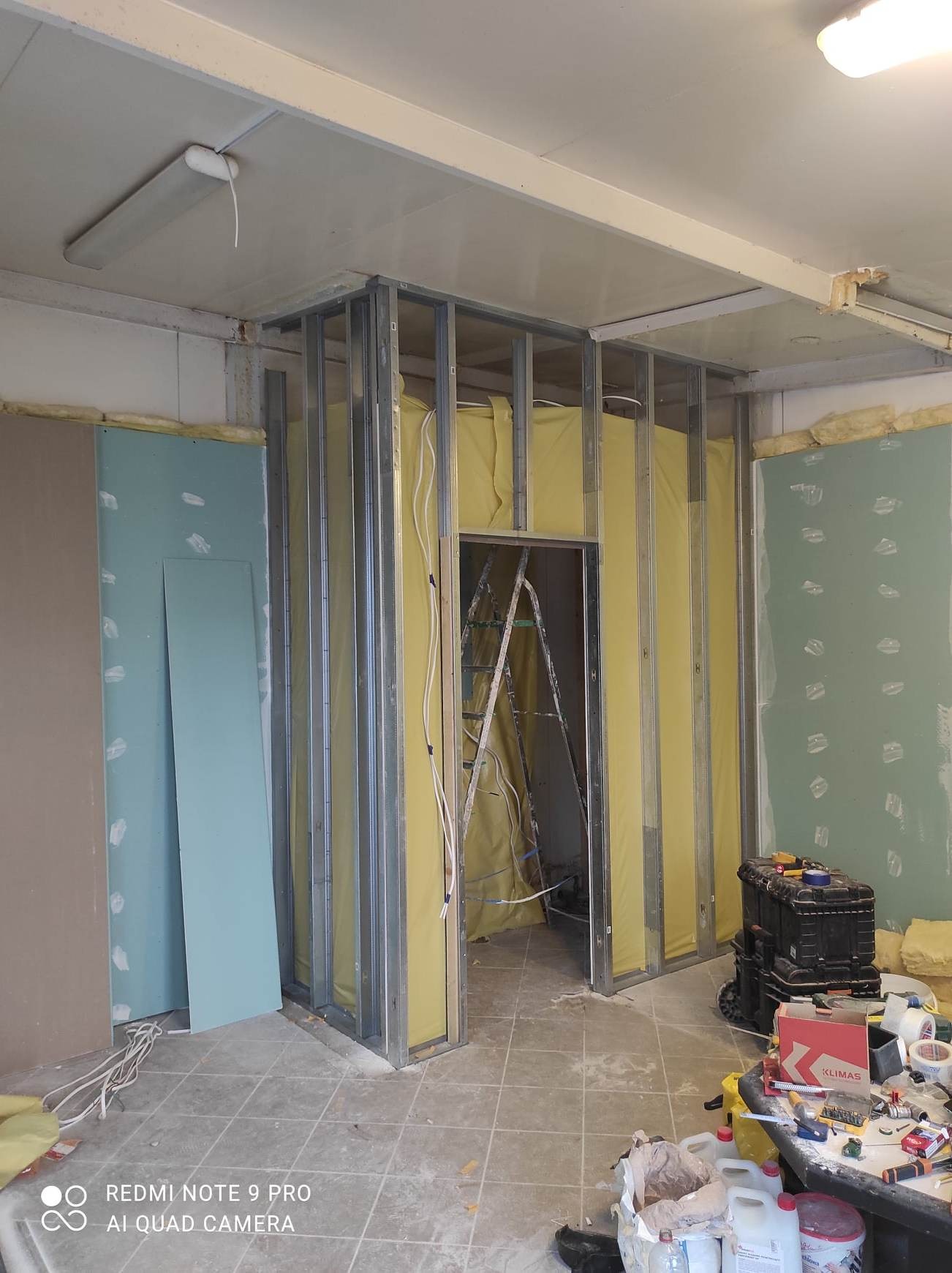 Metalowy szkielet ścianki działowej z wypełnieniem z wełny mineralnej, widoczne przewody elektryczne, płyty gipsowe o różnych odcieniach zieleni oparte o ścianę, drabina w przejściu, narzędzia...