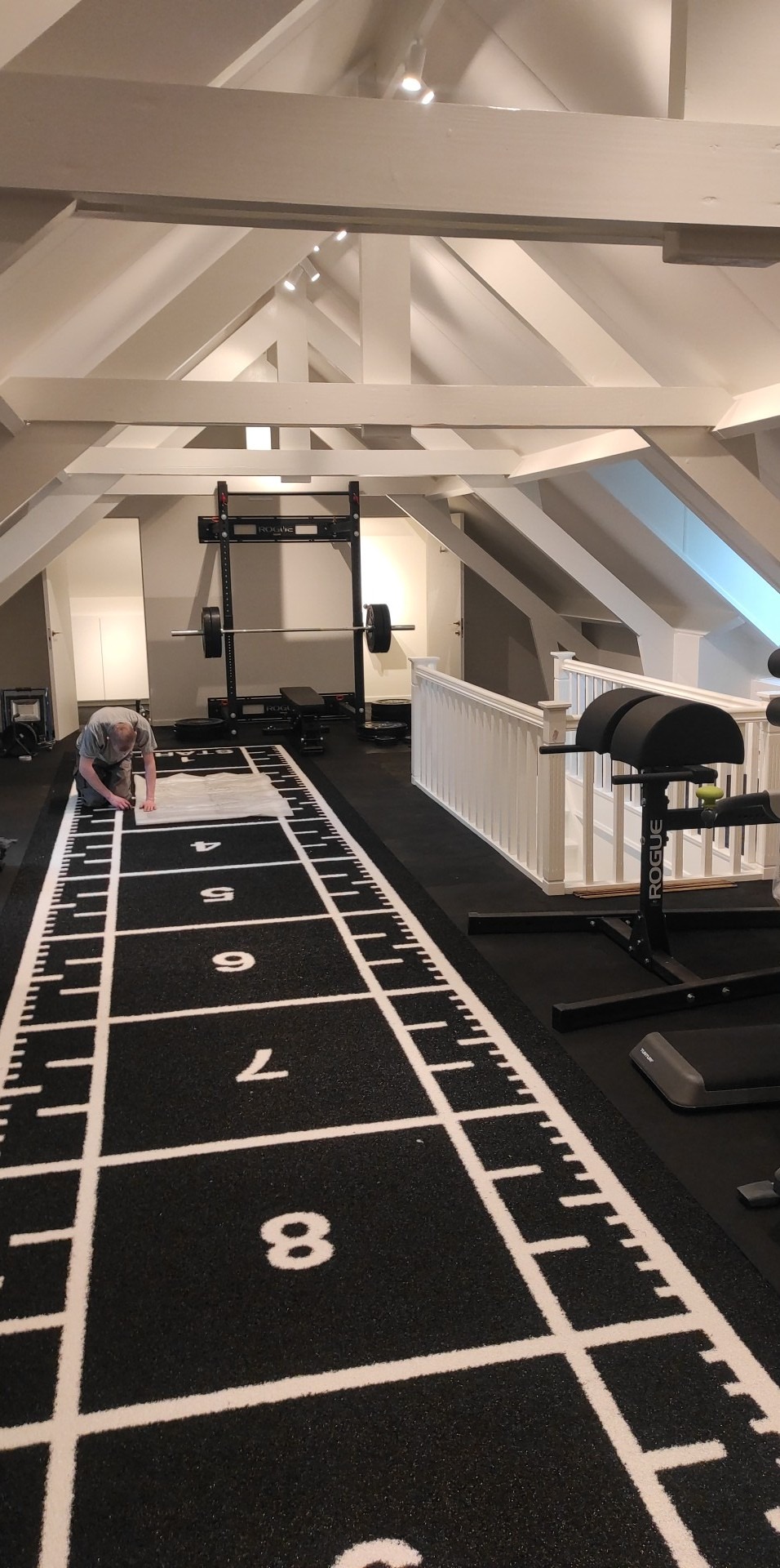 Montaż czarnej, gumowej podłogi w domowej siłowni na poddaszu z numeracją od 1 do 9; widoczny sprzęt sportowy i białe, drewniane belki stropowe.