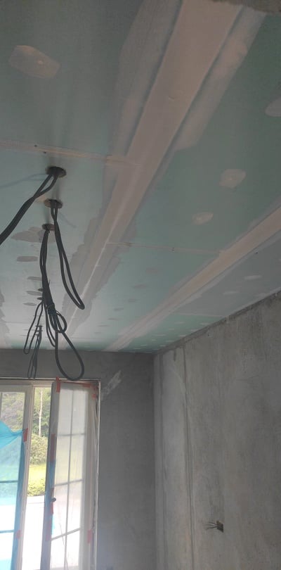 Surowy sufit z zamontowaną płytą gipsowo-kartonową, z widocznymi przewodami elektrycznymi i wstępnie zaszpachlowanymi łączeniami, ściany w stanie surowym przygotowane do dalszych prac...