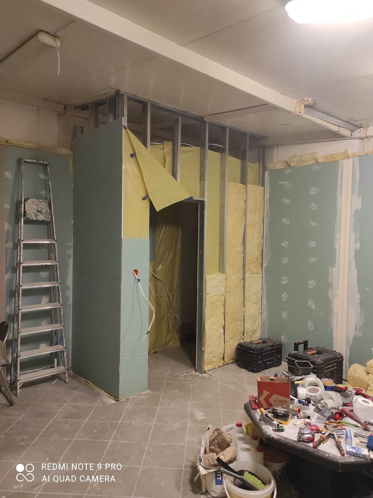 Wnętrze w trakcie remontu: metalowa konstrukcja, izolacja z wełny mineralnej, częściowo zamontowane płyty, drabina oparta o ścianę, narzędzia i materiały budowlane na stole w pierwszym planie.