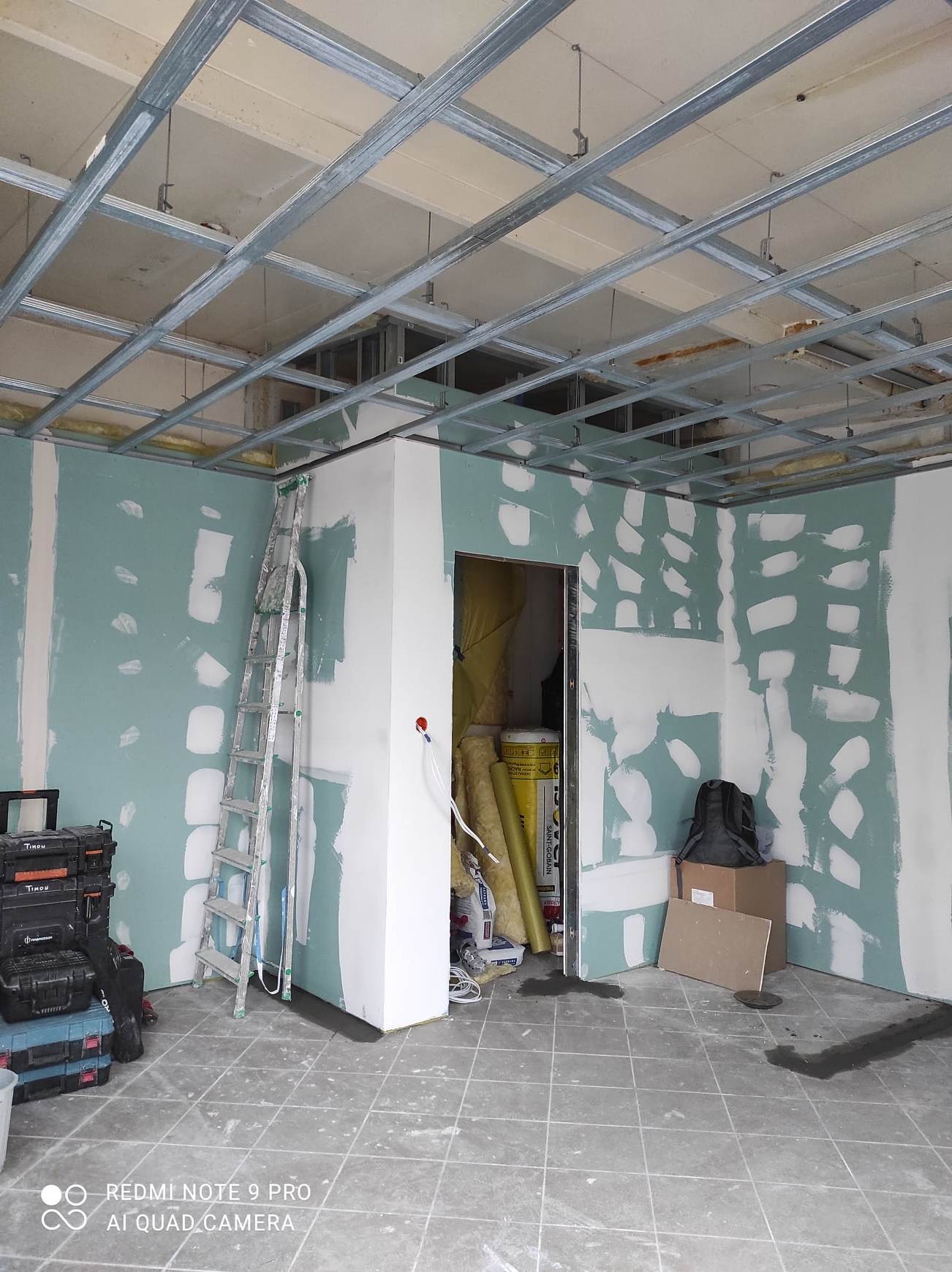 Wnętrze w trakcie prac wykończeniowych: widoczne metalowe profile sufitowe, częściowo oszlifowane ściany z nałożoną gładzią, drabina oparta o ścianę, skrzynki narzędziowe i materiały budowlane...